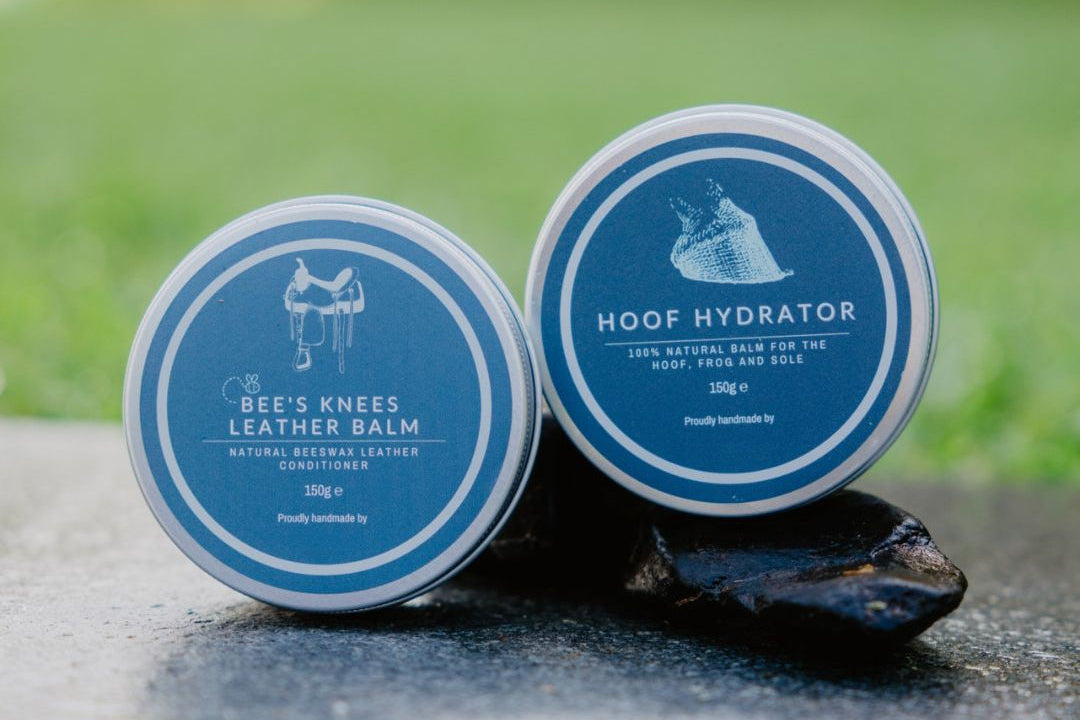 Two containers of 'Bee's Knees Leather Balm' and 'Hoof Hydrator' on a stone surface with a blurred green background.